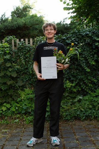 Portrait photo of Felix Deiters with certificate and flowers in front of a green hedge