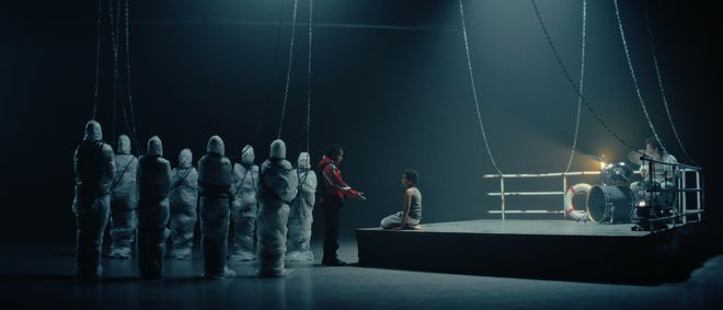 Film still from »DROWNING IS SILENT« by Alexander Knauer: a sea rescuer stands next to a seated boy on a boat-like platform. Several veiled figures are visible on the left, while a drummer sits on the right.
