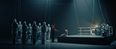 Film still from »DROWNING IS SILENT« by Alexander Knauer: a sea rescuer stands next to a seated boy on a boat-like platform. Several veiled figures are visible on the left, while a drummer sits on the right.