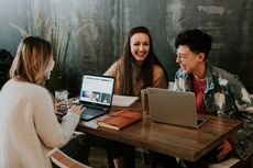 Das Foto zeigt drei junge Frauen, die in einem Café um einen Tisch herum sitzen und sich lachend unterhalten. Auf dem Tisch stehen zwei aufgeklappte Laptops und Getränke.
