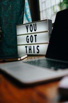 The photo shows a desk with a laptop, a notebook, and a light box with the message: "You got this."