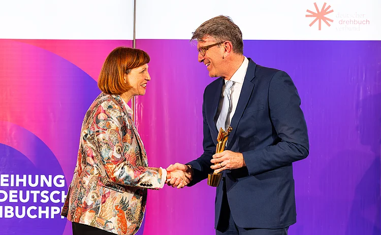 Lena Libertà shakes hands with Germany’s Minister of State for Culture and Media, Wolfram Weimer, on a stage. Wolfram Weimer holds a golden award trophy, and both are smiling in front of a colorful backdrop.