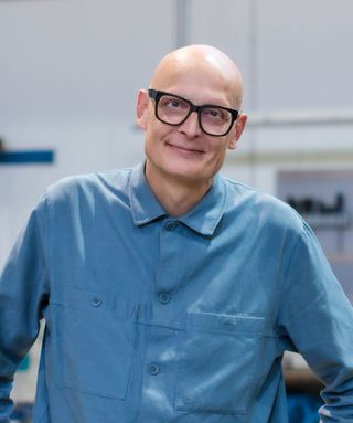 Porträt von Professor Andreas Mühlenberend in der Metallwerkstatt. Er trägt eine hellblaue Jacke und eine schwarze Brille, lehnt sich leicht nach vorn und blickt mit einem leichten Lächeln in die Kamera. Im Hintergrund sind unscharfe Bereiche der Werkstatt zu sehen.