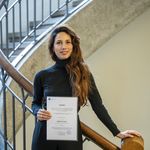 Betül Peker on the stairs in the foyer of the main building, holding her award in front of her.