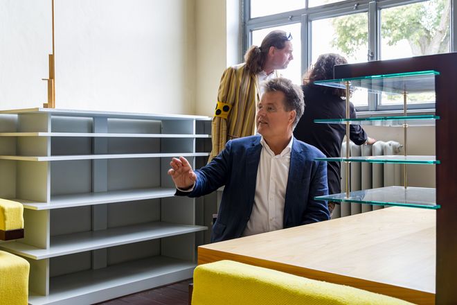 Jürgen Dusel im Gropiuszimmer, er sitzt am Schreibtisch, seine Hand zeigt nach vorne. Im Hintergrund sind Michaela Albrecht und Gunter Seidel zu sehen, die sich unterhalten und aus dem Fenster schauen.
