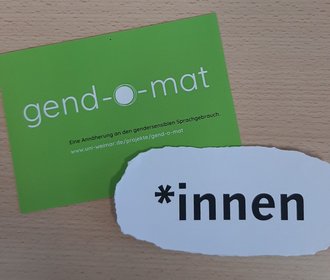 The symbolic image shows a postcard advertisement for the Equal Opportunity Office's »gend-o-mat« as well as a piece of paper with the inscription »*innen«.