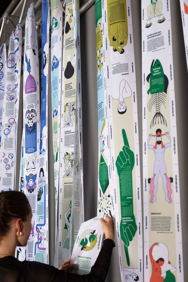 Detail view of a long, colourfully printed fabric strip hanging in several loops from a metal structure. A person holds one section of the strip in their hands.