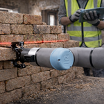 Ein Roboterarm setzt einen Ziegelstein auf eine Ziegelmauer und wird dabei von einer roten Laserlinie ausgerichtet. Im Hintergrund hält eine Person mit Warnweste und Handschuhen ein Tablet in einem teilweise errichteten oder sanierten Gebäude.