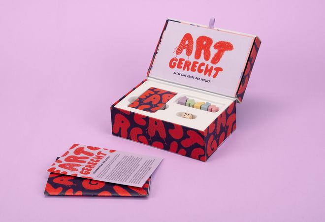 Open game box from »ARTGERECHT« by Anna Buhler: the box contains playing cards, colourful game pieces and a die. A booklet and an information sheet about the board game are placed beside it.
