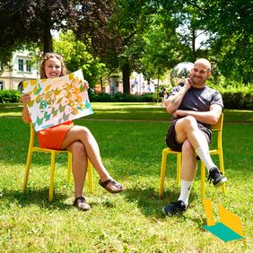 Foto: Nina-Marie Luderer/ Bauhaus-Universität Weimar Frederik Sukop und Beatrice Girth sitzen auf zwei gelben Stühlen in einem Park, sie hält ein Plakat zur summaery in der Hand, er eine Diskokugel