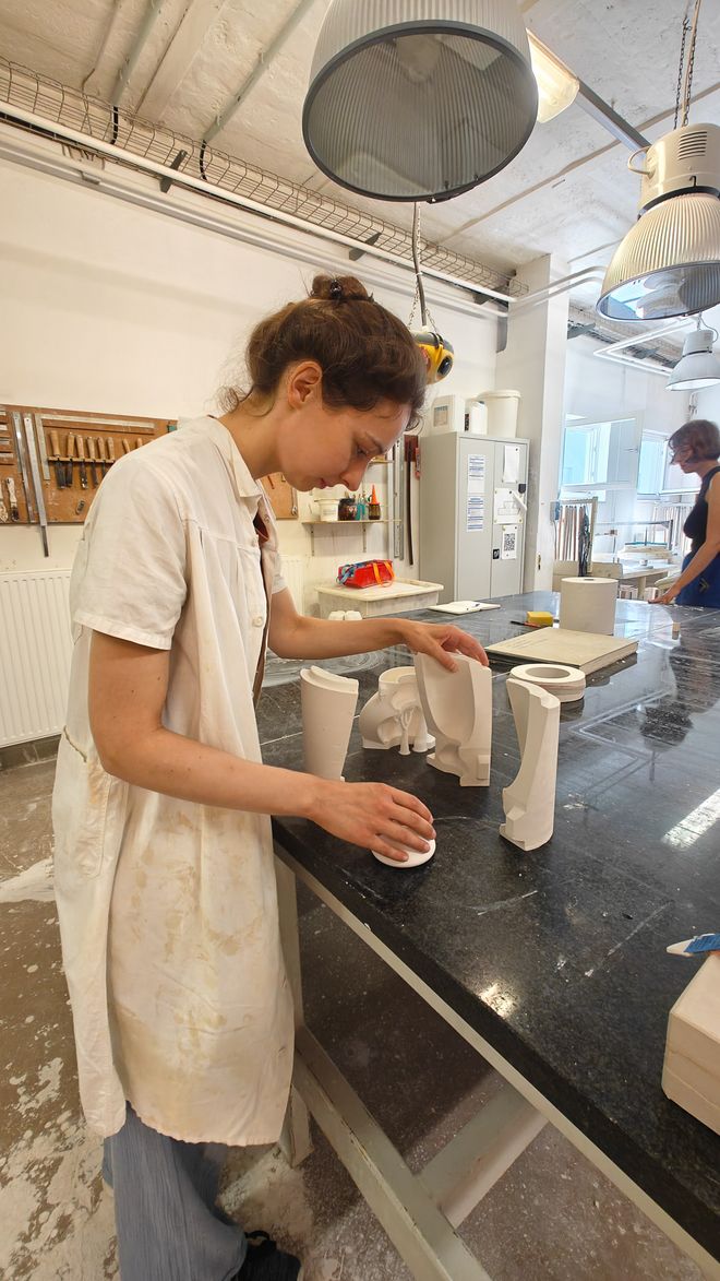 Charlotte setzt einzelne Teile einer weißen Gießform auf einer Werkbank zusammen. Im Hintergrund ist die Gips- und Formenbauwerkstatt zu sehen.