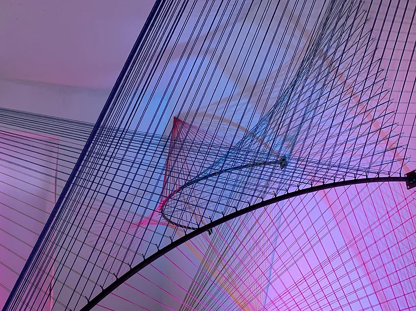 Spatial installation from the “Ein+Ander” project featuring a network of stretched threads and curved metal structures, illuminated in pink, purple and blue.