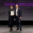Photo: Salar Baygan Marie Kurstjens stands on stage at the German Design Award 2026 ceremony, holding her certificate and trophy as »Newcomer of the Year«; a representative of the event stands beside her, with her name projected prominently in the background.
