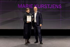 Marie Kurstjens steht bei der Preisverleihung des German Design Award 2026 auf der Bühne und hält ihre Urkunde sowie die Trophäe als »Newcomer of the Year«; neben ihr steht ein Vertreter der Veranstaltung, im Hintergrund ist ihr Name groß projiziert.