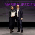 Marie Kurstjens stands on stage at the German Design Award 2026 ceremony, holding her certificate and trophy as »Newcomer of the Year«; a representative of the event stands beside her, with her name projected prominently in the background.