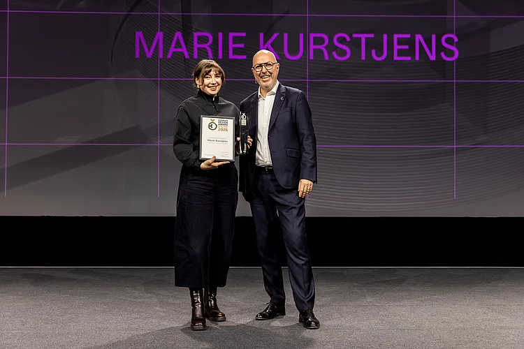 Marie Kurstjens stands on stage at the German Design Award 2026 ceremony, holding her certificate and trophy as »Newcomer of the Year«; a representative of the event stands beside her, with her name projected prominently in the background.