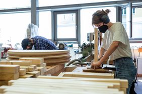 Eine Studentin arbeitet mit Mundschutz in der Holzwerkstatt an einem Möbelstück für die das Foyer der Mensa, im Hintergrund ist ein Student zu sehen. Bauhaus-Universität Weimar, Foto: Thomas Müller