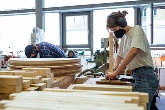 A student wearing a mouth guard works in the wood workshop on a piece of furniture for the foyer of the refectory; a student can be seen in the background. Bauhaus-Universität Weimar, Photo: Thomas Müller