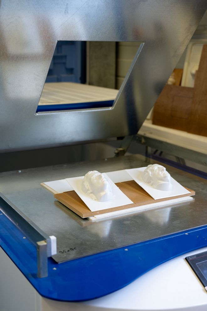 Zwei weiße, kopfförmige Formteile liegen nebeneinander auf einer Trägerplatte in einer Vakuumtiefziehanlage. Links ist ein Schiller-Kopf, rechts ein Goethe-Kopf zu sehen. Darüber befindet sich die metallische Abdeckung der Maschine mit rechteckiger Öffnung.