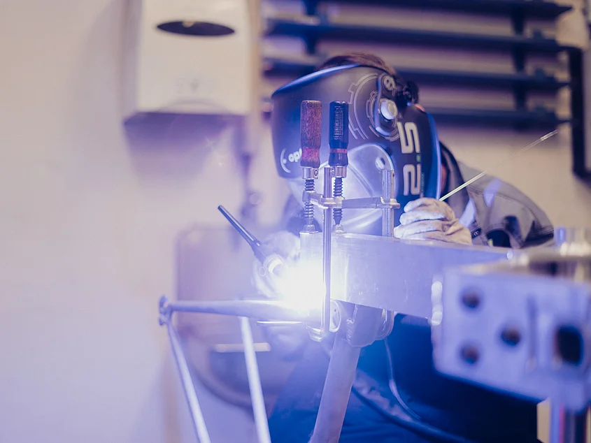 Person mit Schweißhelm arbeitet an einem eingespannten Metallteil; Schraubzwingen fixieren das Werkstück, Schweißlicht ist sichtbar.