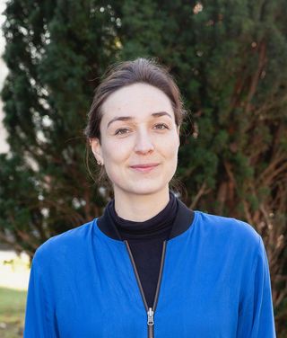 Porträt von Elisa Rufenach-Ruthenberg. Sie trägt eine blaue Jacke über einem schwarzen Oberteil und blickt mit leichtem Lächeln in die Kamera. Im Hintergrund sind unscharfe Bäume zu sehen.