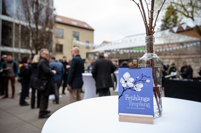 Lila Karte auf der Frühlingsempfang zum Semesterauftakt steht, dahinter stehen Menschen und unterhalten sich 