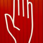 Das Foto zeigt den weißen Umriss einer Hand, die "Achtung!" signalisiert, auf einer roten Mauer.