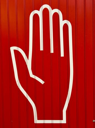 Das Foto zeigt den weißen Umriss einer Hand, die "Achtung!" signalisiert, auf einer roten Mauer.