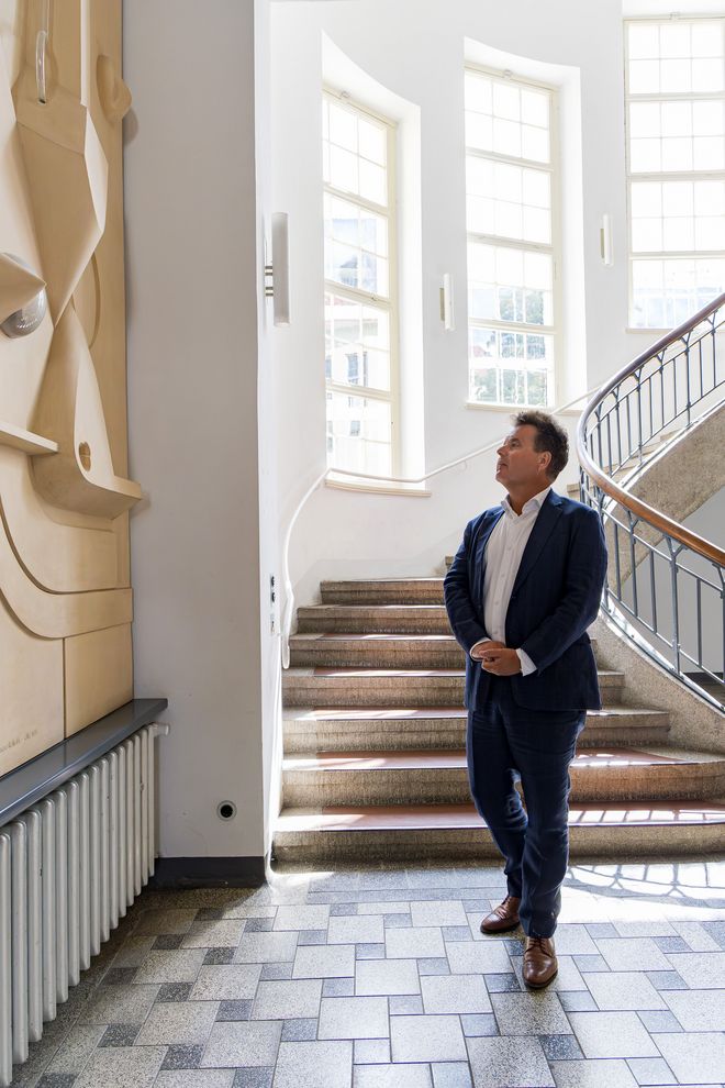 Jürgen Dusel steht im Foyer des Hauptgebäudes, er ist alleine auf dem Foto zu sehen. Er betrachtet sehr interessiert ein Kunstwerk an der Wand: das Wandrelief Konfigurationen von Jost Schmidt von 1923 in einer Nachbildung von Hubert Schiefelbein 1976. Es ist sehr groß, abstrakt geometrisch mit verschiedenen hervorstehenden Formen, hell, aus Stuck und Glas. Schmidt studierte seit 1910 an der Hochschule für Bildende Künste in Weimar und war nach dem 1. Weltkrieg bis 1925 Meisterschüler der Werkstatt für Holzbildhauerei am Bauhaus in Weimar und Dessau. Zur Bauhaus-Ausstellung 1923 erarbeitete er drei abstrakt geometrische Wandreliefs für das Foyer. Eines davon wurde 1976 durch Hubert Schiefelbein rekonstruiert.