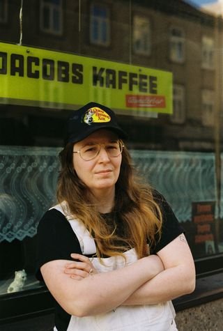 Photo: Lee Everett Thieler Portrait of Teresa Fischer. She wears a black cap, glasses, a black T-shirt and white dungarees. Standing with folded arms in front of a shop window, she looks into the camera.