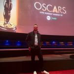 Person steht auf einem roten Teppich in einem großen Kinosaal. Im Hintergrund zeigt eine Leinwand das Oscar-Logo sowie Hinweise zur Liveübertragung der Verleihung.