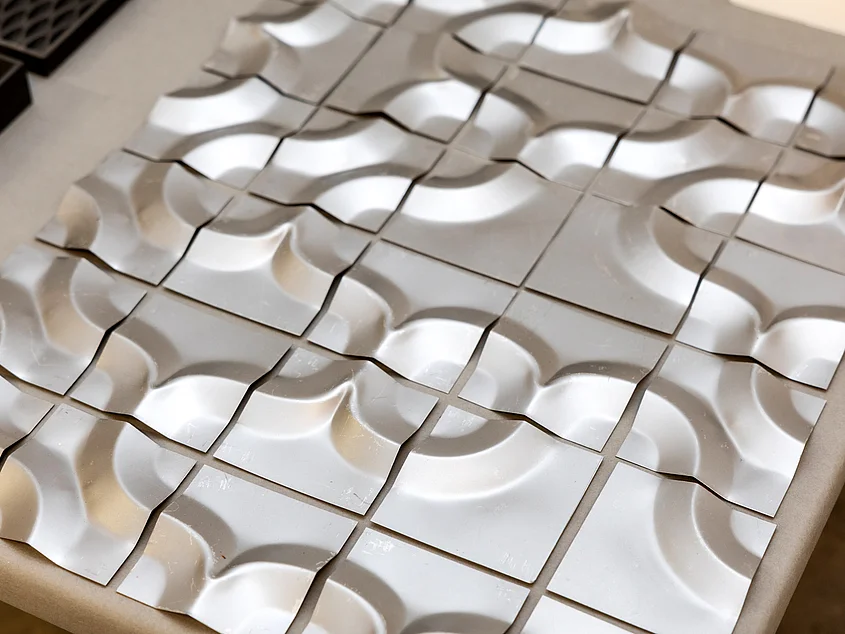 Close-up of a grid-like arrangement of white, square metal elements. The individual pieces feature varying curves and indentations that combine to form a relief-like surface.