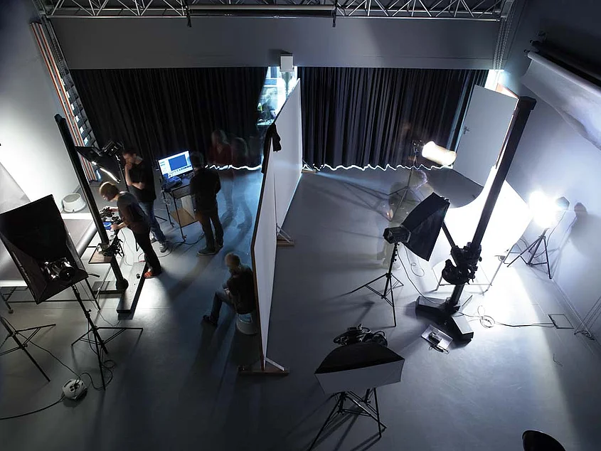 Blick von oben in ein professionell ausgestattetes Fotostudio. Zu sehen sind verschiedene Scheinwerfer, Softboxen, Kameras und Stative sowie mehrere Personen bei der Arbeit. Eine Stellwand teilt den Raum in zwei Arbeitsbereiche.