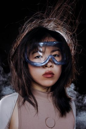 Portrait einer jungen Frau mit Laborbrille, dunklem Haar und neutralem Gesichtsausdruck vor dunklem Hintergrund und leichtem Nebel.