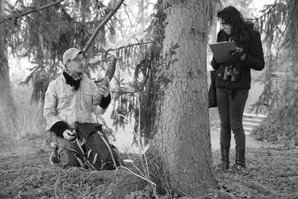 Emanuel Mathias kniet neben einem Baum im Park an der Ilm und hält einen Ast in der Hand; eine Frau steht mit einem Klemmbrett daneben