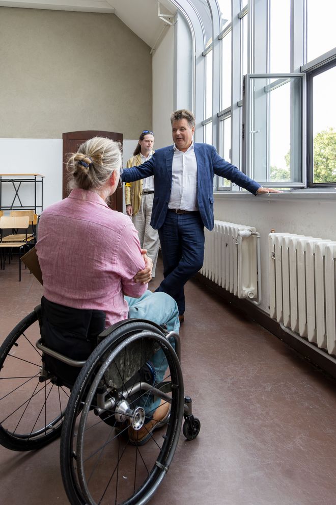 Jürgen Dusel steht vor einem offenen Fenster in einem der Ateliers im Hauptgebäude und unterhält sich mit Martin Ahner, der mit dem Rücken zu uns im Rollstuhl sitzt. Im Hintergrund ist Gunter Seidel zu sehen.