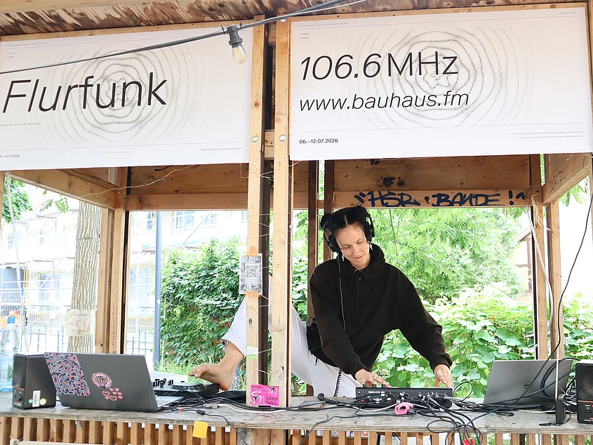 Person wearing headphones operates audio equipment for “Flurfunk” during the summaery. Posters above the improvised broadcasting studio display the frequency 106.6 MHz and bauhaus.fm.