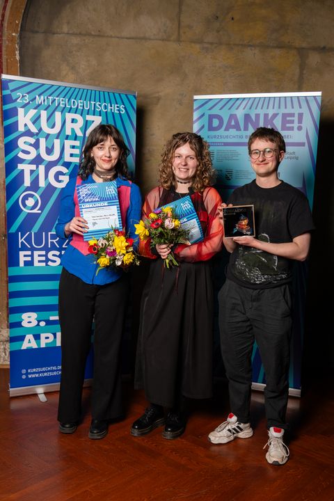 Wir sagen herzlichen Glückwunsch! Tara Thomson, Anya Ryzhkova und Felix Weller stehen bei der Preisverleihung des Kurzsuechtig-Festivals vor Veranstaltungsbannern, halten Urkunden, Blumen und einen Preis in den Händen und blicken in die Kamera.