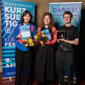 Tara Thomson, Anya Ryzhkova und Felix Weller stehen bei der Preisverleihung des Kurzsuechtig-Festivals vor Veranstaltungsbannern, halten Urkunden, Blumen und einen Preis in den Händen und blicken in die Kamera.