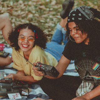 Two young women sit smiling on a meadow. One is holding a radio from the 80s in her hand.