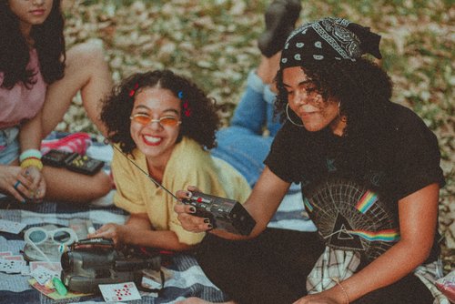 Two young women sit smiling on a meadow. One is holding a radio from the 80s in her hand.