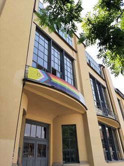 Das Foto zeigt ein Banner mit der »Inter*-inklusiven Progress-Pride-Flagge« am zentralen Balkon des Hauptgebäudes der Bauhaus-Universität Weimar.