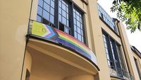 The photo shows a banner with the »Intersex-Inclusive Progress Pride Flag« hanging from the balcony of the Main Building of the Bauhaus-Universität Weimar.