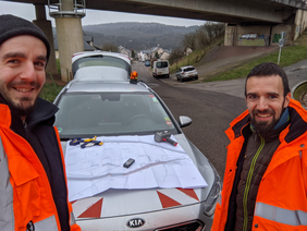 Zwei Männer stehen vor einem Auto auf einer Straße, blicken direkt in die Kamera.