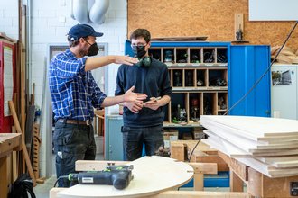 Ein Student spricht mit einem weiteren Studenten in der Holzwerkstatt über ein Möbelstück für die das Foyer der Mensa, beide tragen einen Mundschutz. Bauhaus-Universität Weimar, Foto: Thomas Müller