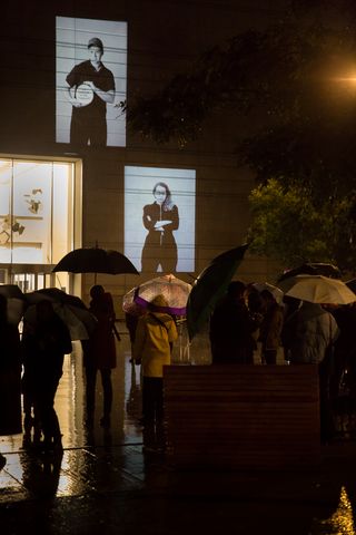 Das Foto zeigt 2 Schwarz-Weiß-Porträts aus Larissa Barths Fotoserie walter ≠ bauhaus, die auf die nächtliche Außenfassade des Bauhaus-Museums Weimar projiziert werden. Passant*innen mit Regenschirmen stehen auf dem Museumsvorplatz und betrachten die Bilder.