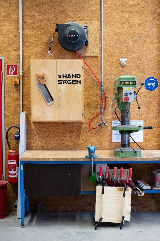 View through a glass window into a woodworking workshop with workbenches, cabinets, and machines. Ceiling lights and extraction systems are visible.