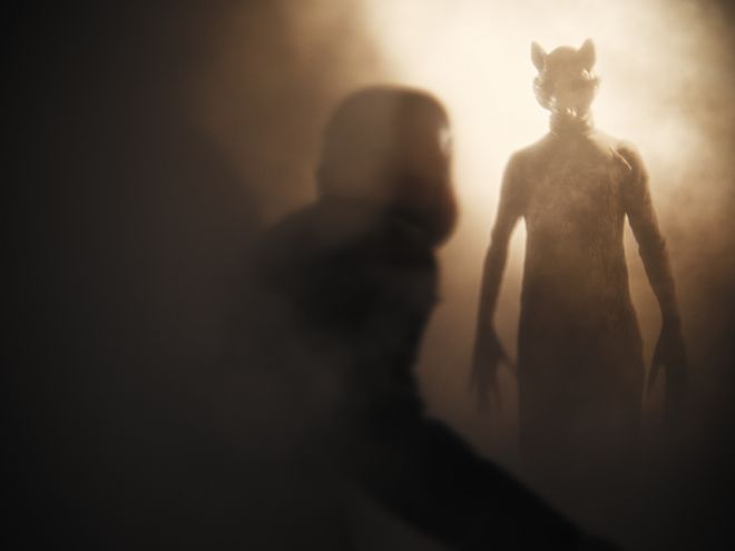 Unscharfe Figur im Vordergrund, im Hintergrund eine hochgewachsene Figur mit tierähnlichem Kopf im Gegenlicht.