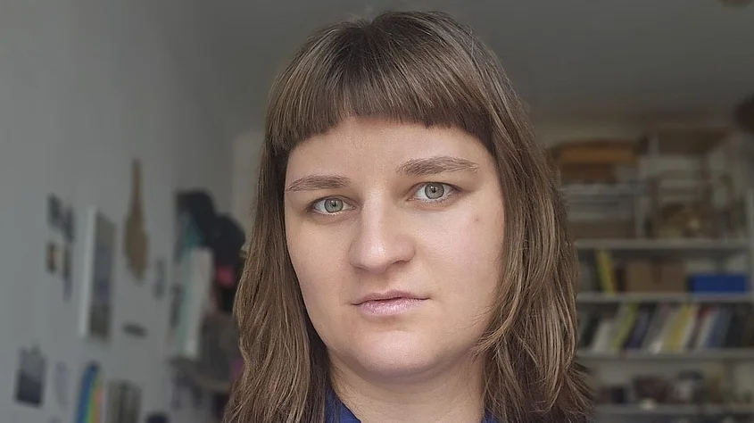 Portrait of Nele Seifert. She has shoulder-length brown hair with bangs and wears gold earrings and a delicate necklace. Set against a softly blurred green background, she looks into the camera with a slight smile.