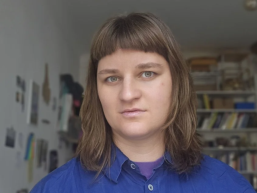 Porträt von Nele Seifert. Sie hat schulterlanges braunes Haar mit Pony und trägt goldfarbene Ohrringe sowie eine feine Halskette. Vor einem weich verlaufenden grünen Hintergrund blickt sie mit leichtem Lächeln in die Kamera.